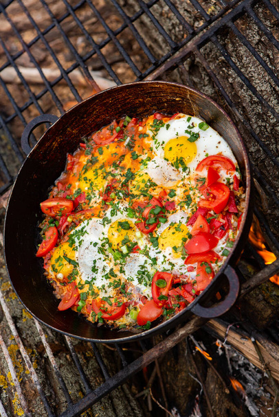Eggs prepared with vegetables on a frying pan on the fire in the forest. Cooking scrambled eggs over an open fire. Breakfast in nature. Tourist food. - Photo, Image