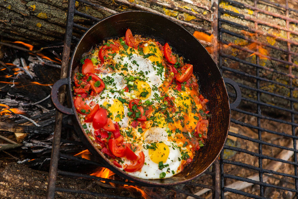 Eieren bereid met groenten op een koekenpan op het vuur in het bos. Roerei bakken boven een open vuur. Ontbijt in de natuur. Toeristisch voedsel. - Foto, afbeelding