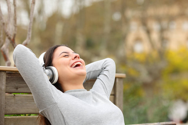 Happy woman listening to music wearing headphones relaxing sitting on a bench in a park - Foto, immagini