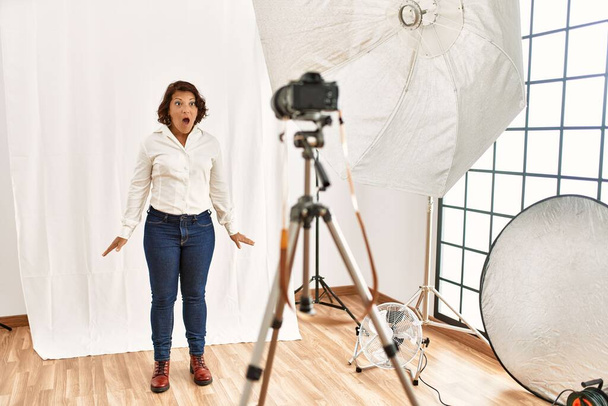Middle age hispanic woman posing as model at photography studio scared and amazed with open mouth for surprise, disbelief face  - Photo, Image
