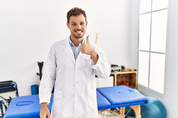 Handsome young man working at pain recovery clinic doing happy thumbs up gesture with hand. approving expression looking at the camera showing success.  - Zdjęcie, obraz