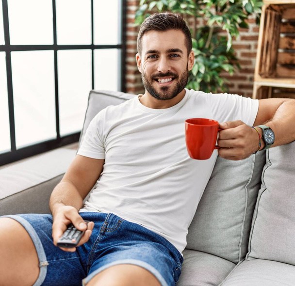 Νεαρός Ισπανός που πίνει καφέ βλέποντας τηλεόραση στο σπίτι - Φωτογραφία, εικόνα