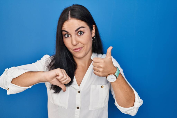 Young hispanic woman standing over blue background doing thumbs up and down, disagreement and agreement expression. crazy conflict  - 写真・画像