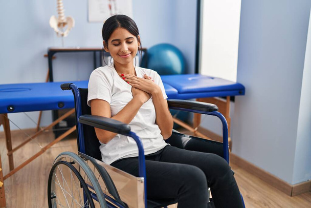 Junge hispanische Frau, die im Rollstuhl in der Physiotherapie-Klinik sitzt, lächelt mit den Händen auf der Brust, die Augen geschlossen mit dankbarer Geste im Gesicht. Gesundheitskonzept.  - Foto, Bild