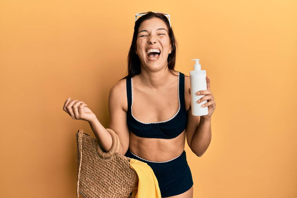 Nuori latino nainen yllään bikinit ja hattu tilalla kesän paju käsilaukku ja aurinkovoidetta hymyilee ja nauraa kovaa ääneen, koska hauska hullu vitsi.  - Valokuva, kuva