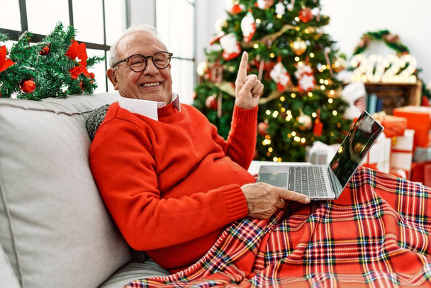 Старший мужчина с седыми волосами с помощью ноутбука сидит на рождественской елке улыбаясь счастливо указывая рукой и пальцем в сторону  - Фото, изображение