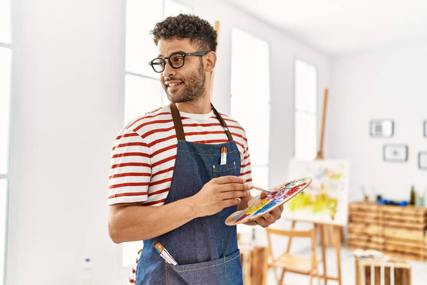 Mladý arabský umělec muž usměvavý šťastný drží štětec a paletu v uměleckém studiu. - Fotografie, Obrázek
