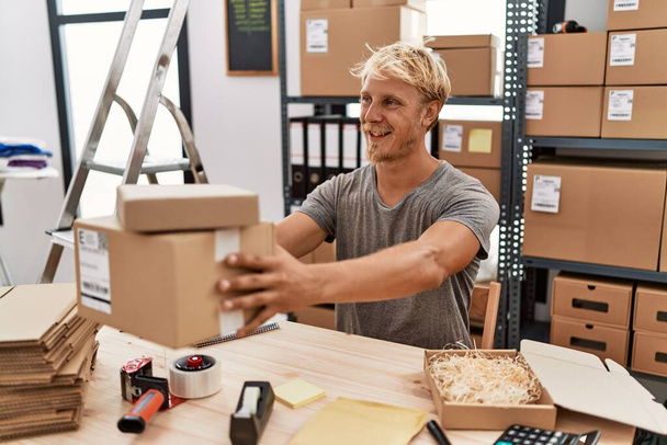 Young caucasian man smiling confident holding package at warehouse - Fotoğraf, Görsel