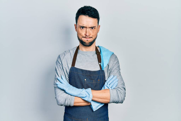 Young hispanic man wearing apron with arms crossed puffing cheeks with funny face. mouth inflated with air, catching air.  - Photo, Image