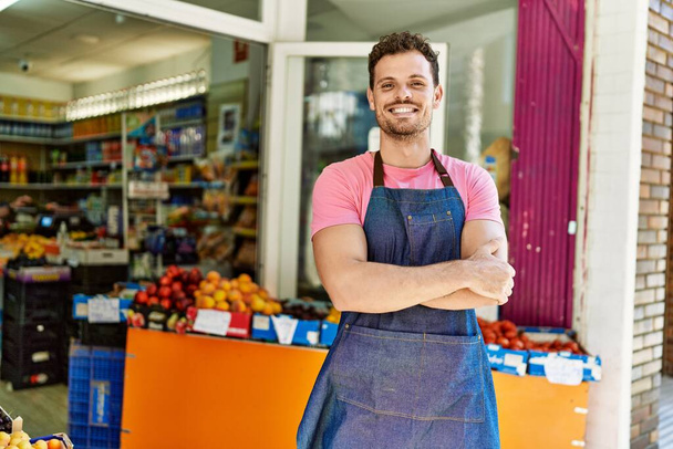 Mladý hispánský dělník v zástěře stojící se zkříženýma rukama v obchodě s ovocem. - Fotografie, Obrázek