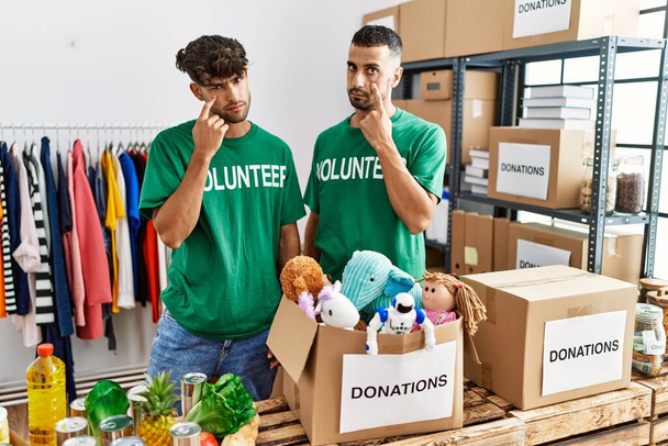 Young gay couple wearing volunteer t shirt at donations stand pointing to the eye watching you gesture, suspicious expression  - Foto, imagen