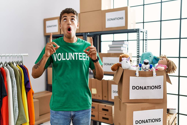 Jonge knappe Spaanse man draagt vrijwilliger t-shirt bij donaties staan verbaasd en verrast omhoog te kijken en wijzen met vingers en opgeheven armen.  - Foto, afbeelding