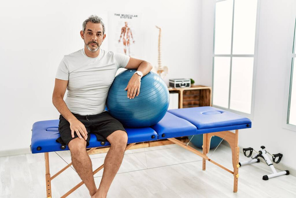 Middle age hispanic man at pain recovery clinic holding pilates ball relaxed with serious expression on face. simple and natural looking at the camera.  - Photo, Image