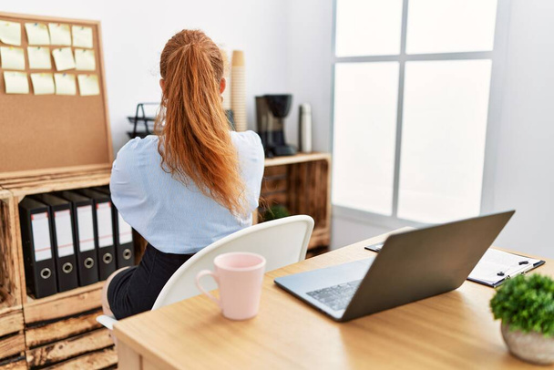 Jeune rousse femme travaillant au bureau en utilisant ordinateur portable debout vers l'arrière regardant loin avec les bras croisés  - Photo, image