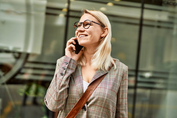 Νεαρή ξανθιά επιχειρηματίας χαμογελώντας ευτυχισμένη μιλώντας στο smartphone στην πόλη. - Φωτογραφία, εικόνα