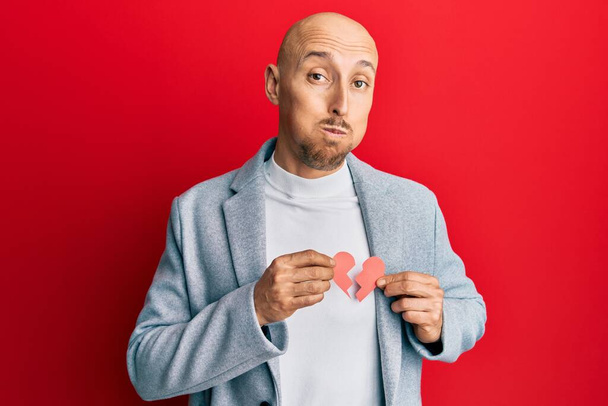 Bald man with beard holding broken heart paper shape puffing cheeks with funny face. mouth inflated with air, catching air.  - Foto, immagini