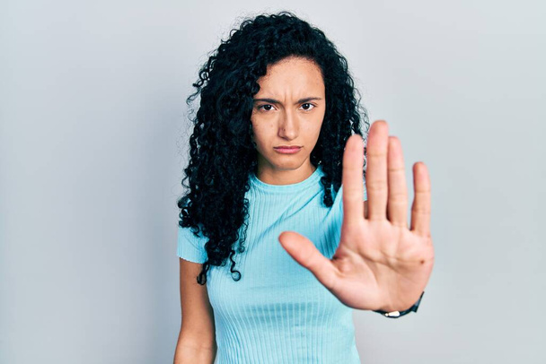 Jonge Spaanse vrouw met krullend haar draagt casual blauw t-shirt doen stoppen met zingen met palm van de hand. waarschuwingsuitdrukking met negatief en ernstig gebaar op het gezicht.  - Foto, afbeelding