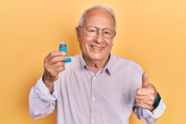 Älterer Mann mit grauen Haaren, der einen Asthma-Inhalator in der Hand hält, lächelt glücklich und positiv, Daumen hoch und Zeichen der Anerkennung  - Foto, Bild