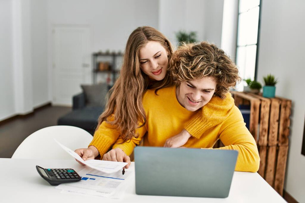 Genç beyaz çift, evde dizüstü bilgisayar kullanarak aile muhasebesi yaparak birbirlerine sarılıyorlar. - Fotoğraf, Görsel