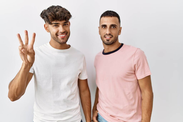 Νεαρό γκέι ζευγάρι στέκεται πάνω από απομονωμένο φόντο που δείχνει και δείχνει προς τα πάνω με τα δάχτυλα νούμερο τρία, ενώ χαμογελά αυτοπεποίθηση και χαρούμενος.  - Φωτογραφία, εικόνα
