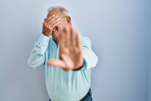 Starszy mężczyzna z siwymi włosami stojący nad niebieskim tłem, zakrywający oczy dłońmi i wykonujący gest zatrzymania ze smutkiem i strachem. wstydliwa i negatywna koncepcja.  - Zdjęcie, obraz