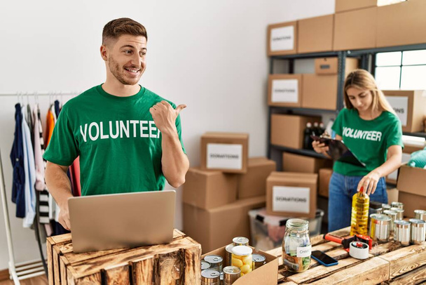 Young man and woman wearing volunteer t shirt at donations stand pointing thumb up to the side smiling happy with open mouth  - Foto, Bild