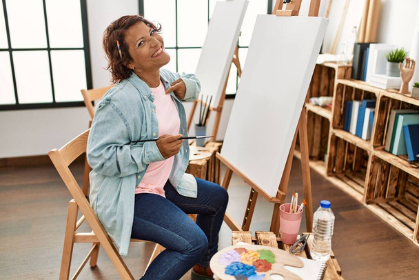 Mulher hispânica de meia idade no estúdio de arte pintura sobre tela apontando dedo para um auto sorrindo feliz e orgulhoso  - Foto, Imagem
