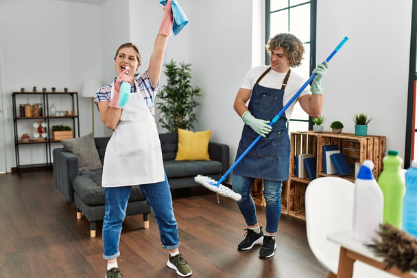 Young caucasian couple cleaning house. Standing with smile on face singing using mop and diffuser as a microphone. - Foto, Imagem