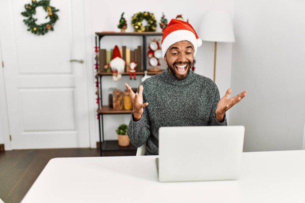 Młody Latynos z brodą w świątecznym kapeluszu z laptopem świętującym zwycięstwo szczęśliwym uśmiechem i wyrazem zwycięzcy z podniesionymi rękami  - Zdjęcie, obraz