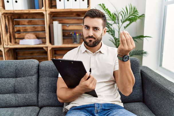 Handsome hispanic man holding clipboard working at psychology clinic doing italian gesture with hand and fingers confident expression  - Foto, Imagen