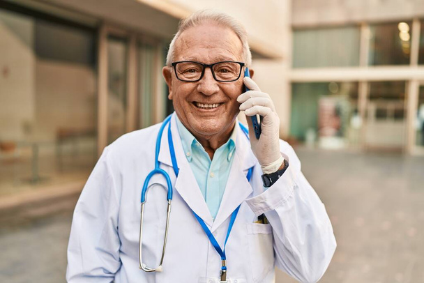 Senior man wearing doctor uniform talking on the smartphone at street - Φωτογραφία, εικόνα
