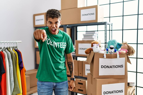 Young handsome hispanic man wearing volunteer t shirt at donations stand pointing displeased and frustrated to the camera, angry and furious with you  - Fotografie, Obrázek