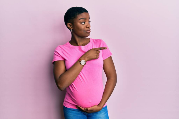 Young african american woman expecting a baby, touching pregnant belly pointing with hand finger to the side showing advertisement, serious and calm face  - Valokuva, kuva
