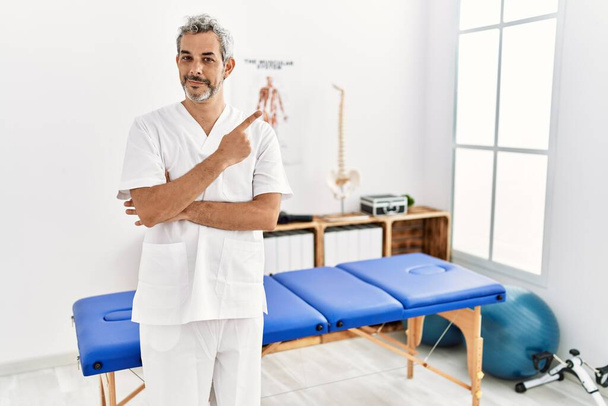Middle age hispanic therapist man working at pain recovery clinic pointing with hand finger to the side showing advertisement, serious and calm face  - Photo, image