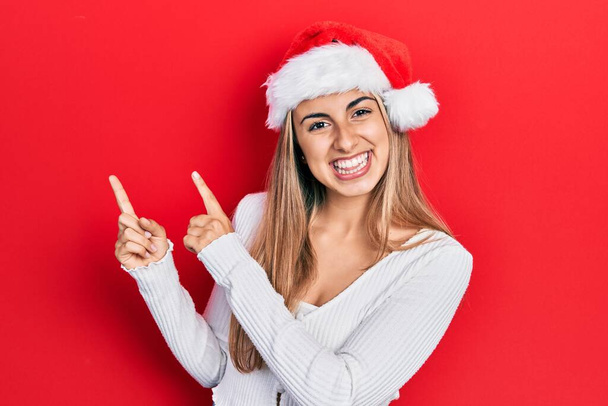Güzel İspanyol kadın Noel şapkası takıyor gülümsüyor ve kameraya bakıyor iki eliyle ve parmaklarıyla kenarı işaret ediyor..  - Fotoğraf, Görsel