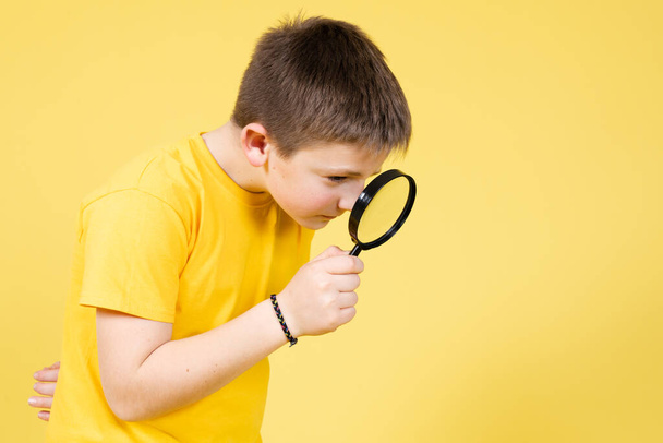 Lindo chico jugando a ser detective y buscando pistas usando una lupa aislada sobre fondo amarillo - Foto, imagen