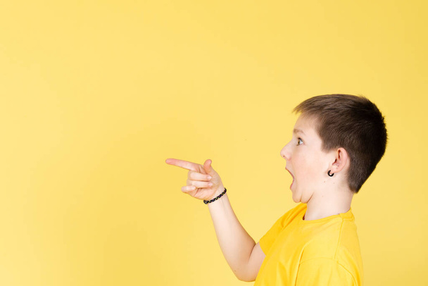Şaşırmış çocuk sarı arka planda izole edilmiş kopya alanına bakıyor ve işaret ediyor. - Fotoğraf, Görsel
