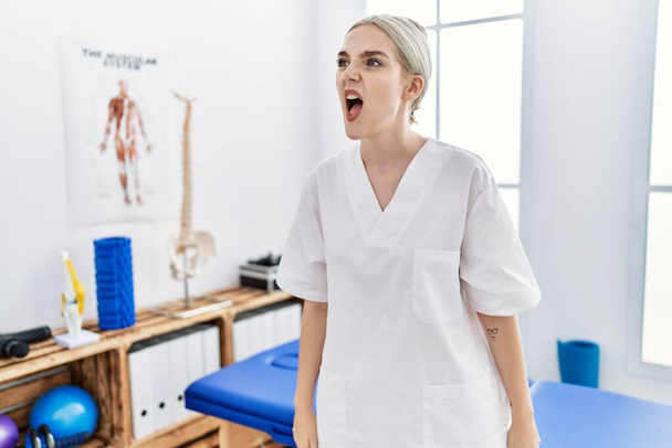 Young caucasian woman working at pain recovery clinic angry and mad screaming frustrated and furious, shouting with anger. rage and aggressive concept.  - 写真・画像