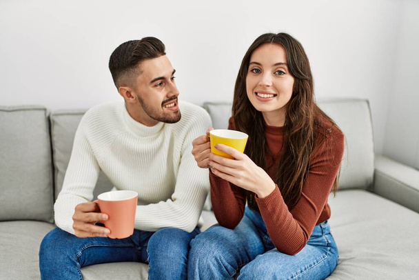 Νεαρό ζευγάρι ισπανόφωνων πίνει καφέ καθισμένο στον καναπέ στο σπίτι. - Φωτογραφία, εικόνα