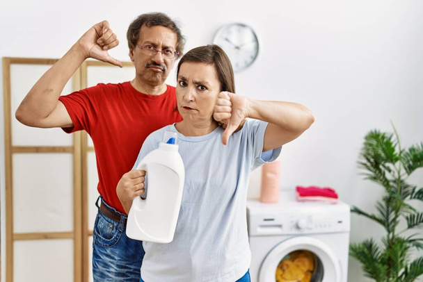 Międzyrasowa para w średnim wieku robi pranie trzymając butelkę po detergentach z rozgniewaną twarzą, negatywne oznaki niechęci do kciuków w dół, koncepcja odrzucenia  - Zdjęcie, obraz