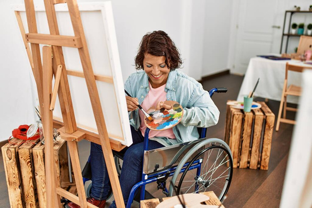 Латиноамериканская художница-инвалид средних лет рисует сидя на инвалидной коляске в художественной студии. - Фото, изображение
