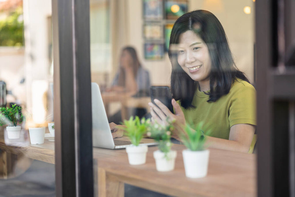Азиатская деловая женщина работает из дома с мобильного телефона и технологии ноутбука в счастье чувствовать себя за столом рядом со стеклом в современном кафе или офисе, владелец бизнеса и предприниматель концепции - Фото, изображение