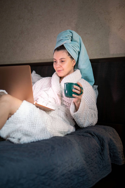 kobieta leżąca w łóżku z mokrą głową pokrytą ręcznikiem pracującym na laptopie pijącym herbatę - Zdjęcie, obraz