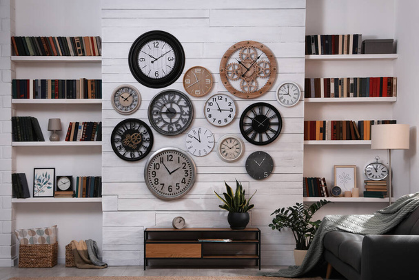 Elegante habitación interior con mesa de consola, cómodo sofá y colección de diferentes relojes en la pared blanca - Foto, Imagen