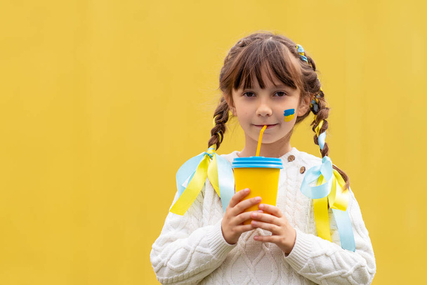 A little Ukrainian girl with two pigtails and yellow and blue ribbons drinks a hot drink from a glass of patriotic colors like the Ukrainian flag. Stop the war in Ukraine. Volunteer Help for Refugees. High quality photo - Foto, imagen