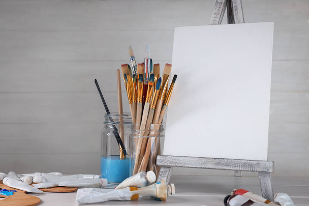 Many brushes, easel with blank canvas and paints on white table - 写真・画像
