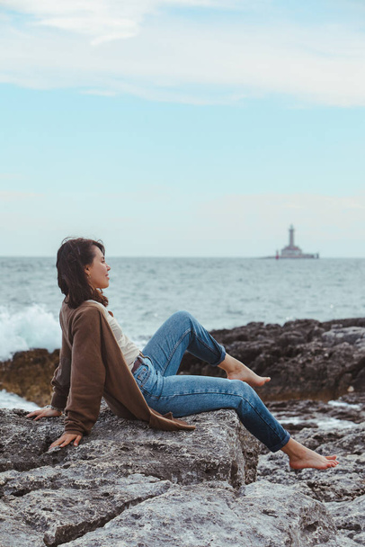 woman sitting by rocky sea beach in wet jeans lighthouse on background. windy weather. summer vacation. carefree concept - Photo, Image