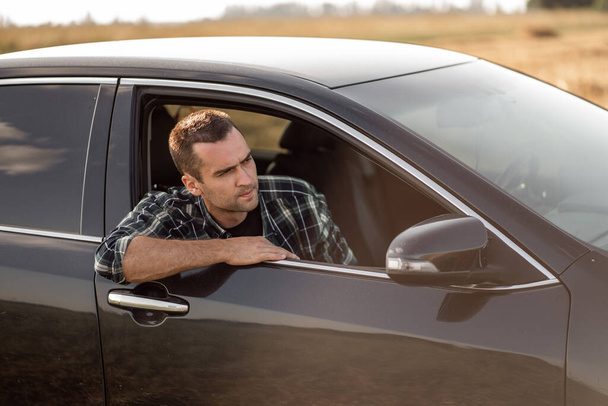 hombre guapo con cerdas cortas en una camisa a cuadros mira por la ventana del coche - Foto, Imagen