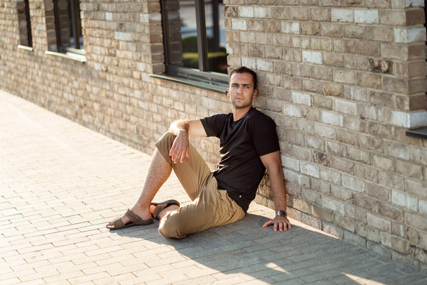 handsome man with short bristles in a black T-shirt on a background of a brick wall sitting on the floor - Φωτογραφία, εικόνα