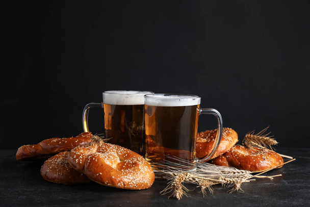 Tasty pretzels, glasses of beer and wheat spikes on black table against dark background, space for text - Photo, image